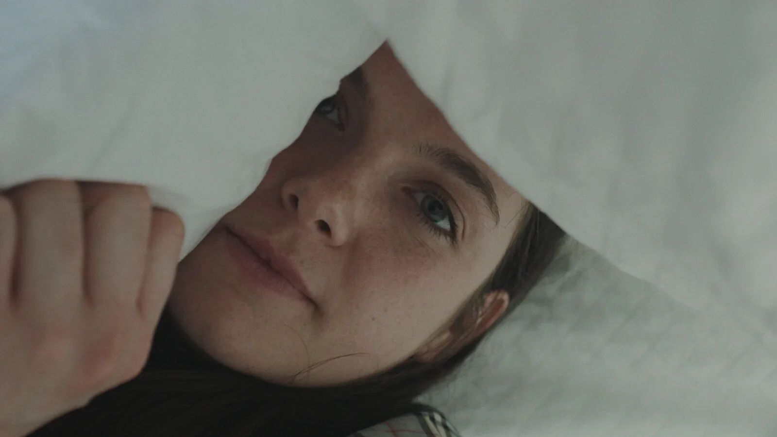 A young girl with freckles and blue eyes is looking through a hole in a sheet of paper, with her face partially obscured.