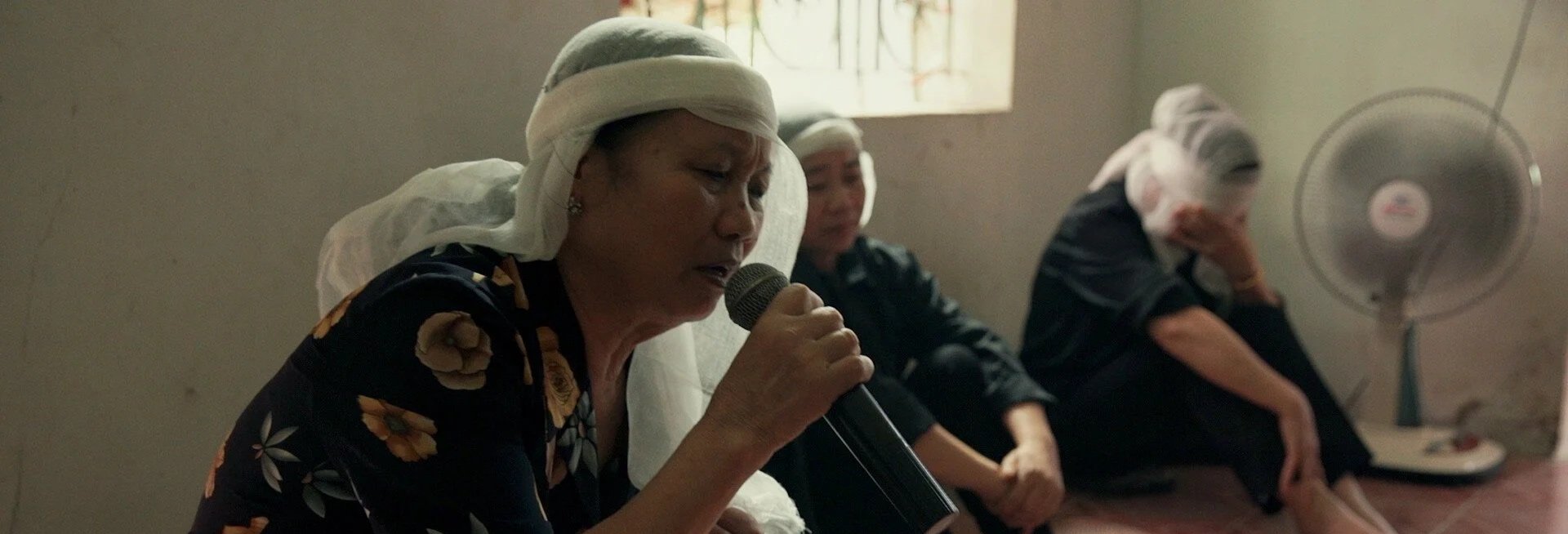 Three women sitting in a room, one woman is speaking into a microphone while wearing a white cap, and the other two women appear to be resting or grieving, with one sitting with her head resting on her hand near a fan.