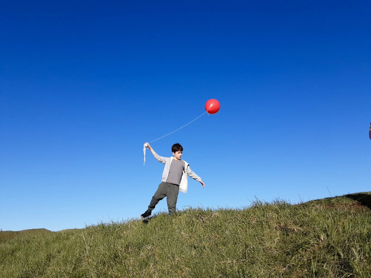 Child holding a red balloon on a string standing on a grassy hill under a clear blue sky.