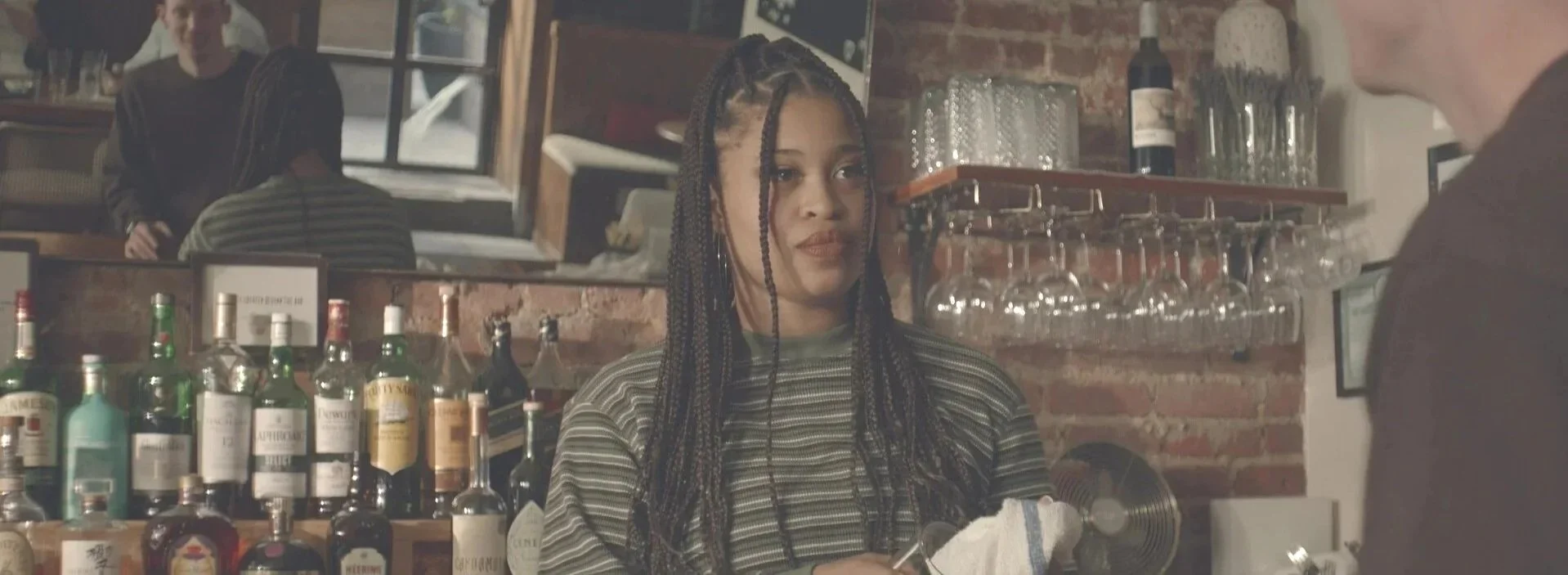 A young woman with braided hair sits at a bar counter, looking at a bartender who is partially visible. The background features a brick wall, bottles of alcohol, and hanging wine glasses.