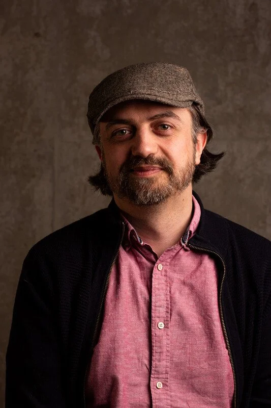 Portrait of a man with a beard, wearing a brown flat cap, pink button-up shirt, and black jacket, standing against a neutral background.