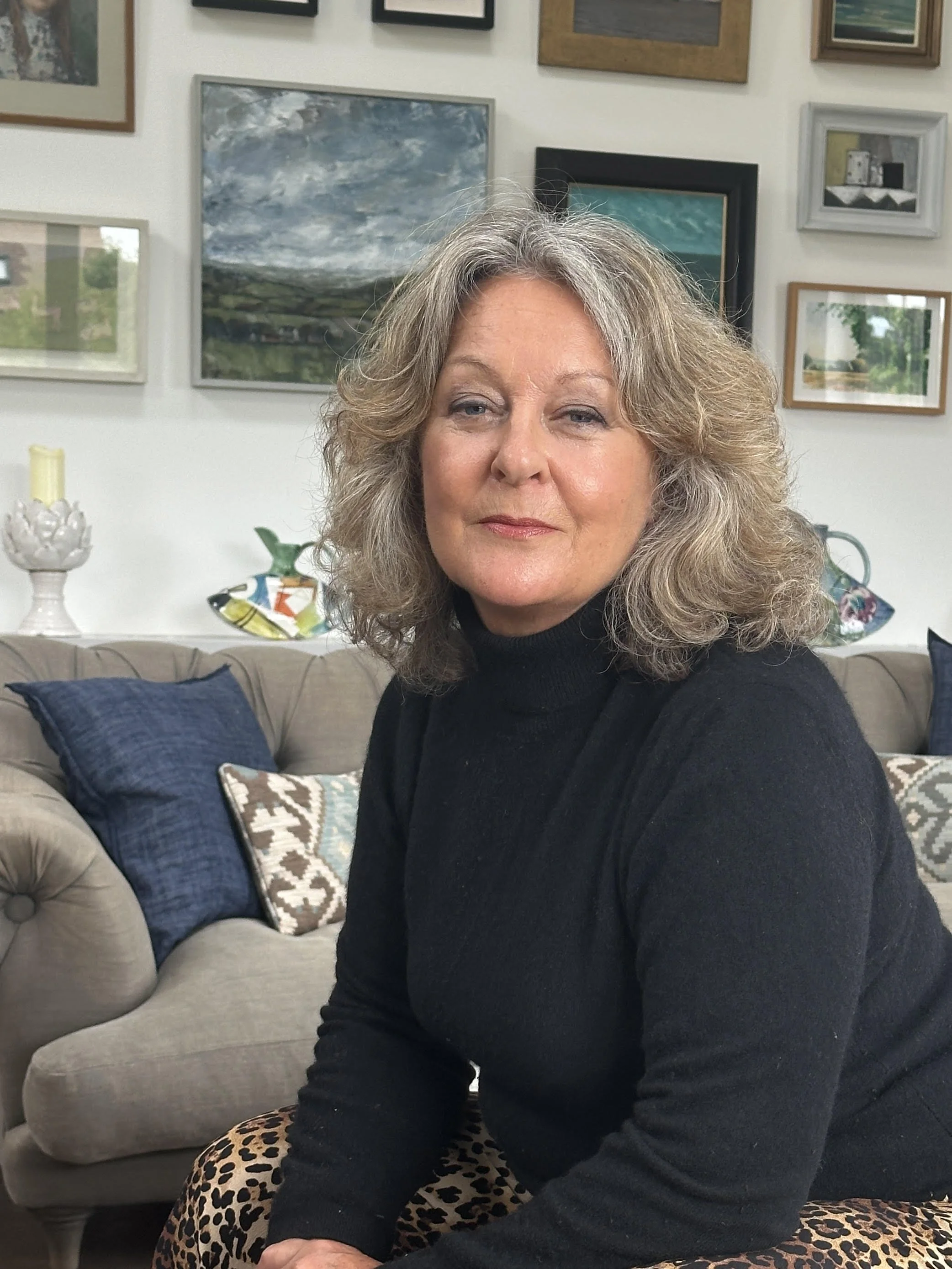 A woman with curly gray hair and light skin, wearing a black turtleneck, sitting on a couch in a living room with framed artwork on the wall behind her.