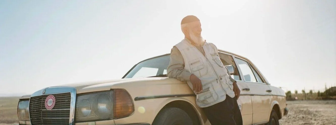 An older man with a beard standing next to a cream-colored vintage car in a desert landscape during daytime.