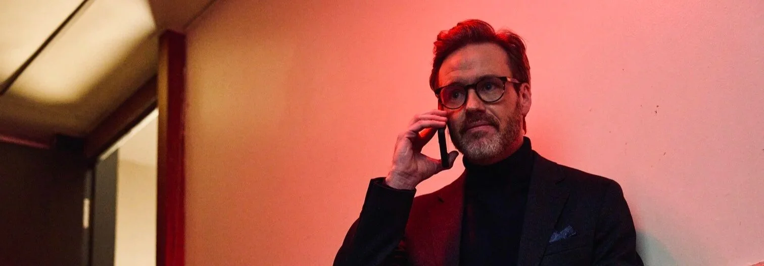 Man with glasses and beard in a black suit talking on a phone against a pinkish wall
