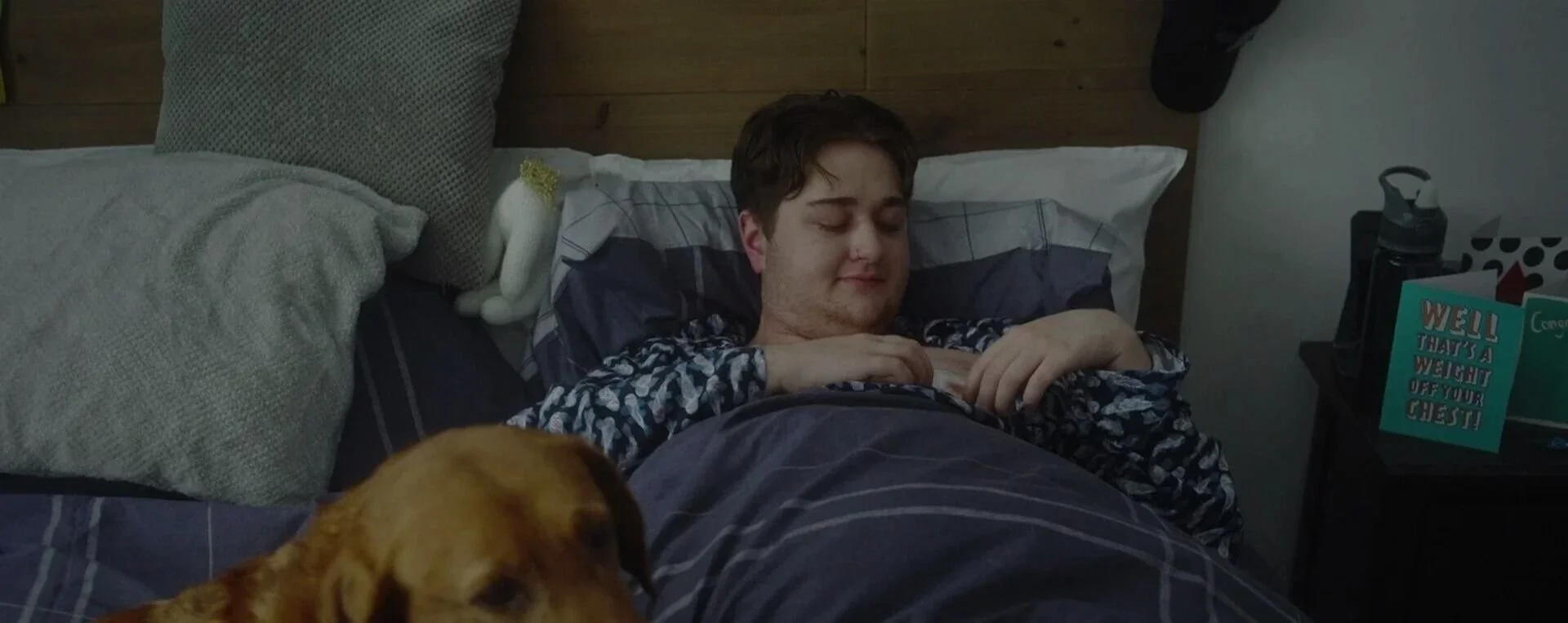 Young man lying in bed with eyes closed, a dog resting near his feet, bedside table with a water bottle, a framed card reading 'Well that's a weight off your chest,' and pillows against a wooden headboard.