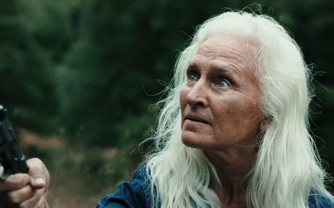 Older woman with long white hair holding a handgun, looking upwards with a serious expression outdoors.