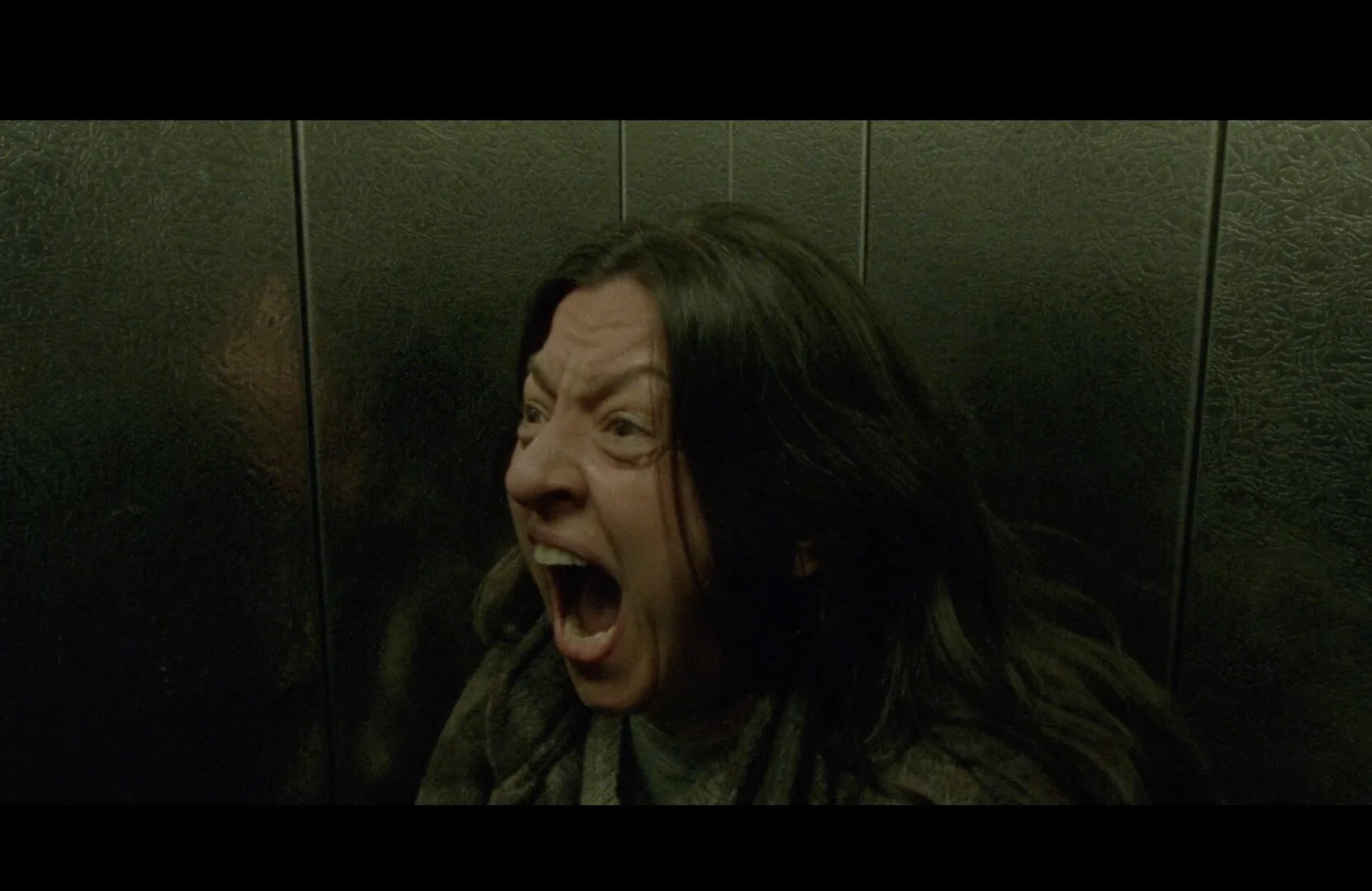 A woman with long dark hair appears to be shouting or screaming with an emotional expression, seated against a dark wall.