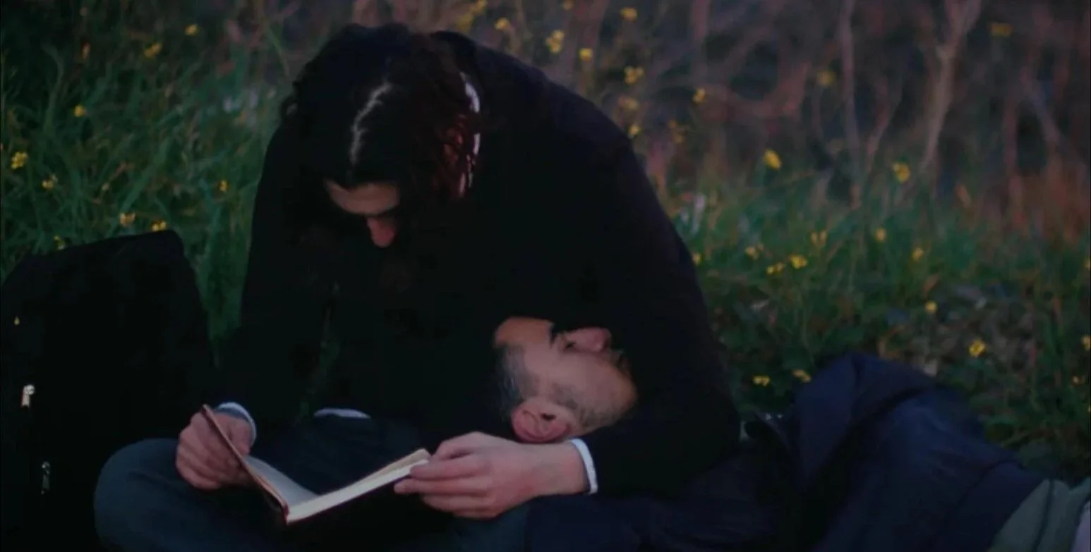 A woman is kneeling over a man lying on the ground outdoors at dusk, with her hand on his face, and they are close together. The man is holding a book and is resting with his eyes closed, while the woman appears to be comforting him.