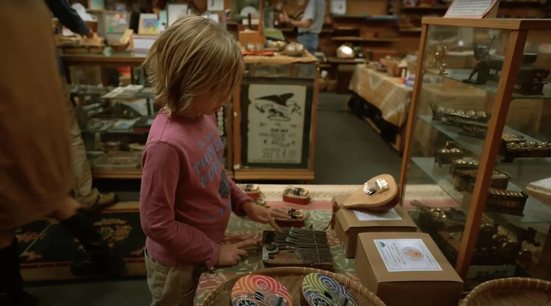 A young boy with long blonde hair wearing a red shirt shopping for utensils in a store.