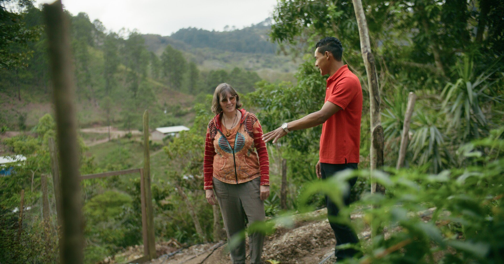 A woman with sunglasses and a colorful jacket smiling while talking to a man in a red polo shirt in a lush, green outdoor area with trees and hills in the background.