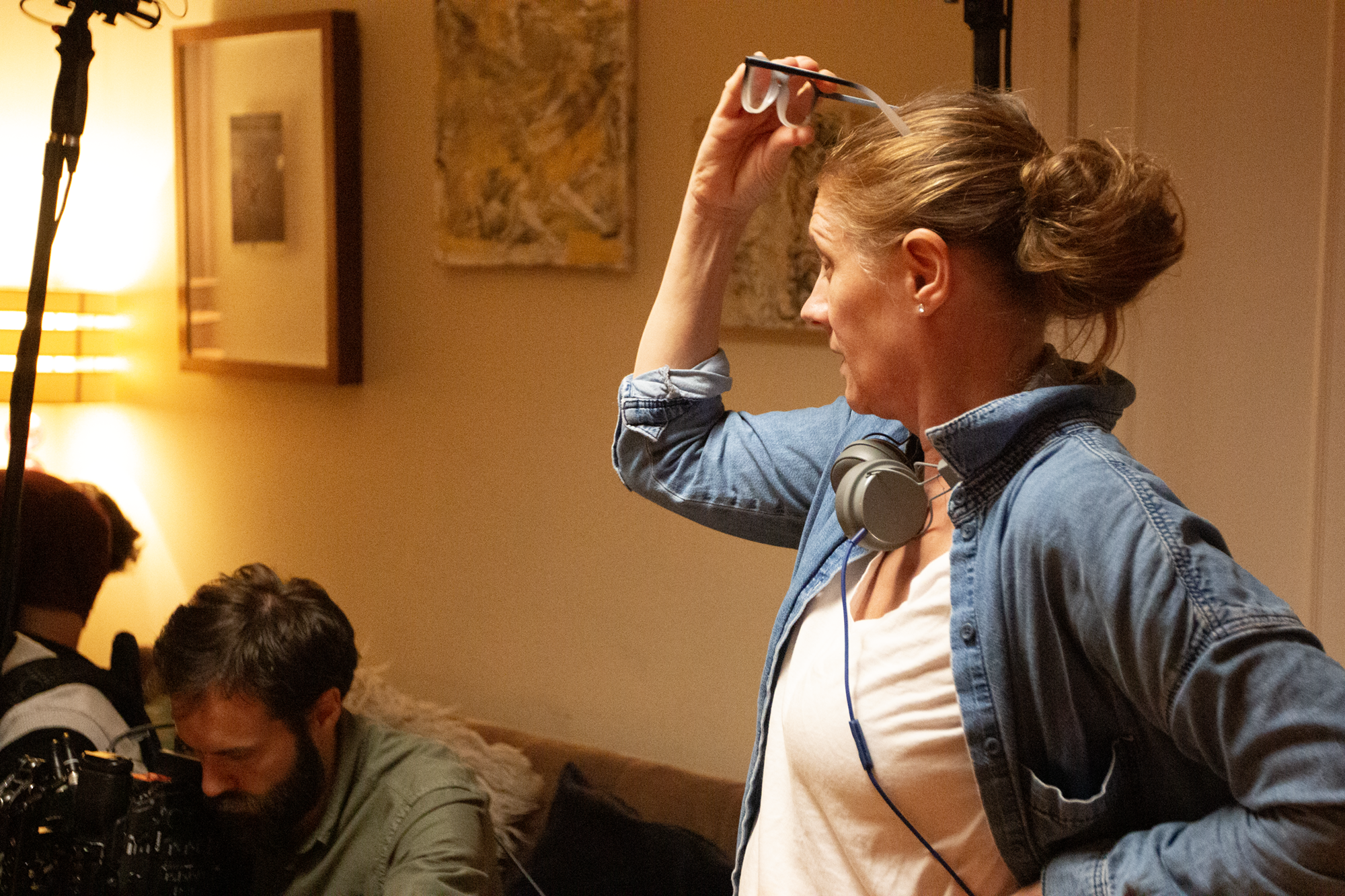 A woman with a denim shirt and headphones around her neck is holding glasses on her head, standing in a room with warm lighting and abstract paintings on the wall, while a man with a beard and a gray shirt is looking down at equipment, possibly on a film or video production set.