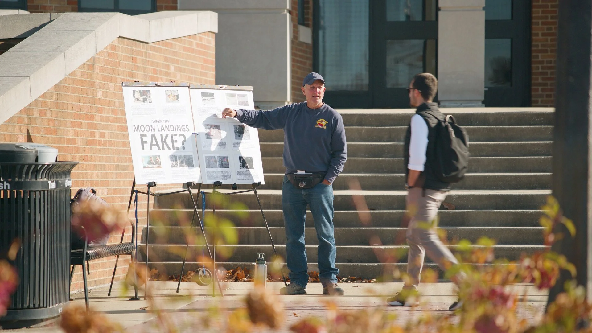 Man in a gray sweatshirt with a logo standing outside, pointing at a presentation board titled 'Were the Moon Landings Fake?' in conversation with a student carrying a backpack, on a city sidewalk near steps and a building.