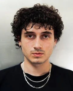 Close-up portrait of a young man with curly dark hair, fair skin, and dark eyes, wearing multiple silver necklaces, against a plain light background.