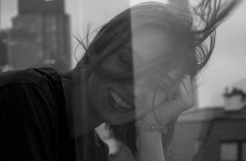 A woman smiling and laughing, resting her head on her hand in an outdoor urban setting, with wind blowing her hair.