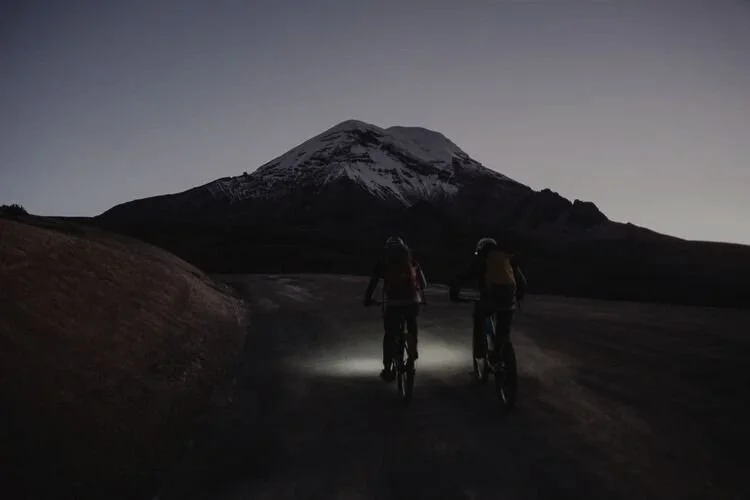 Two people riding mountain bikes on a dirt trail at dusk with a snow-capped mountain in the background.