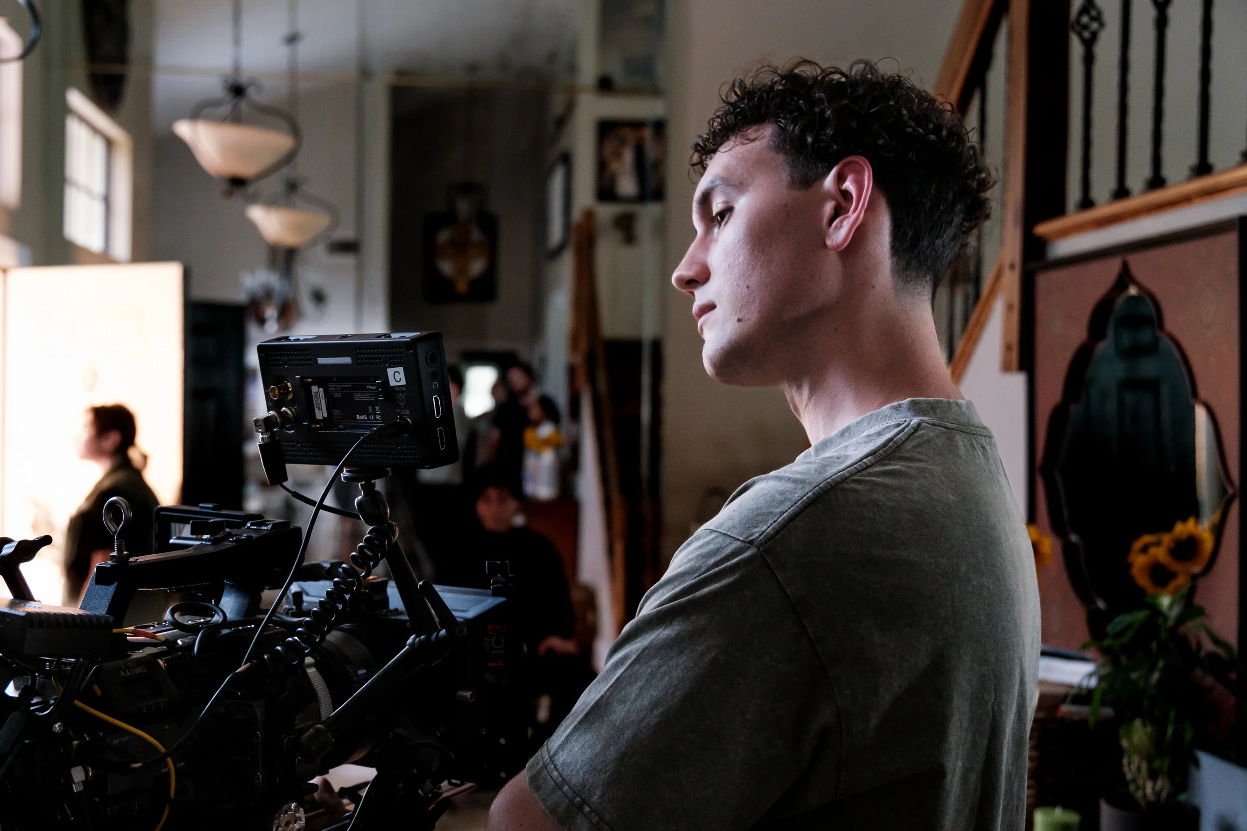 A man with curly hair and a gray t-shirt operating a camera on a film set or studio, with a woman blurred in the background indoors.