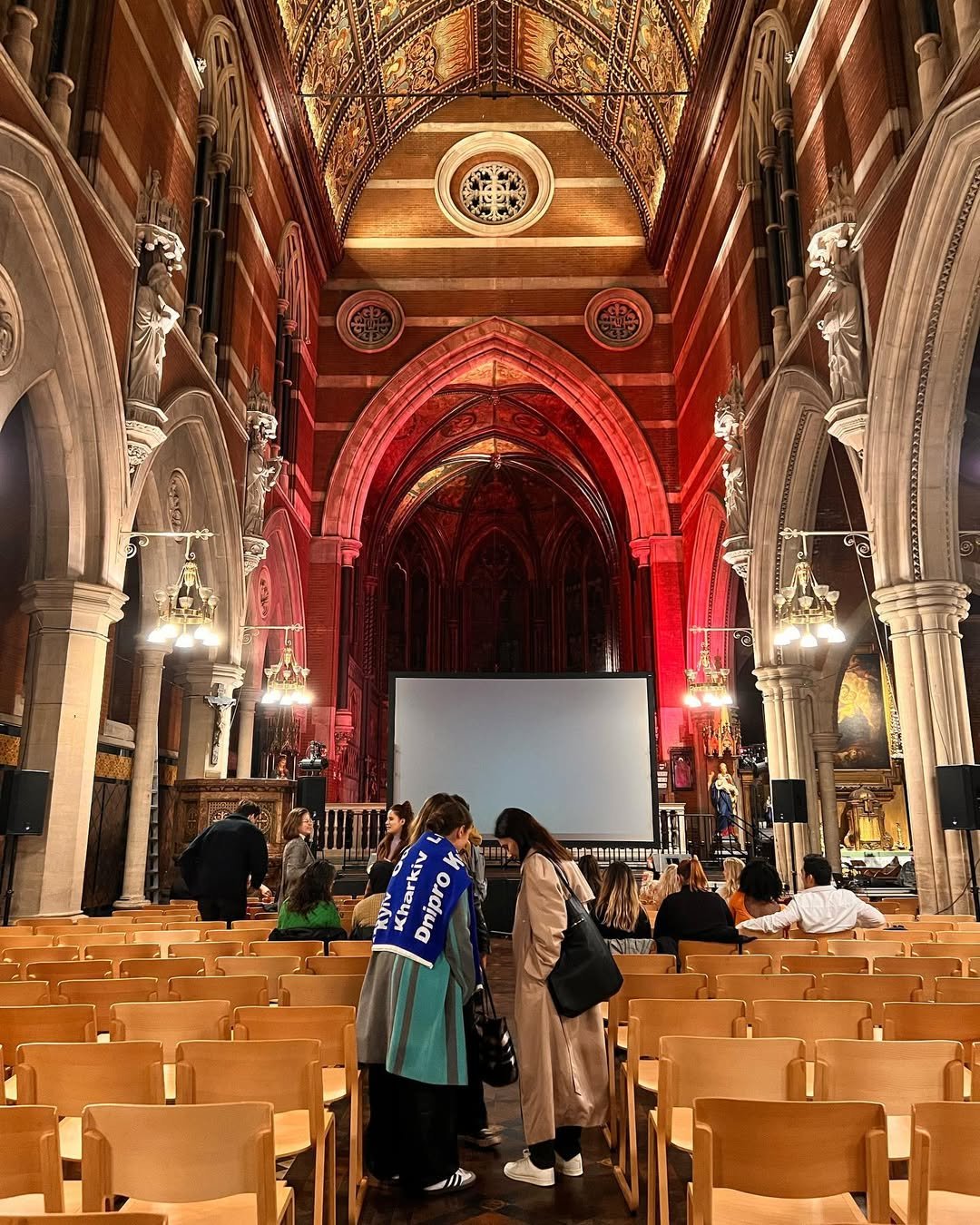 Inside church with ornate architecture, statues, and a large blank screen at the front, people are seated and standing, preparing for an event or presentation.