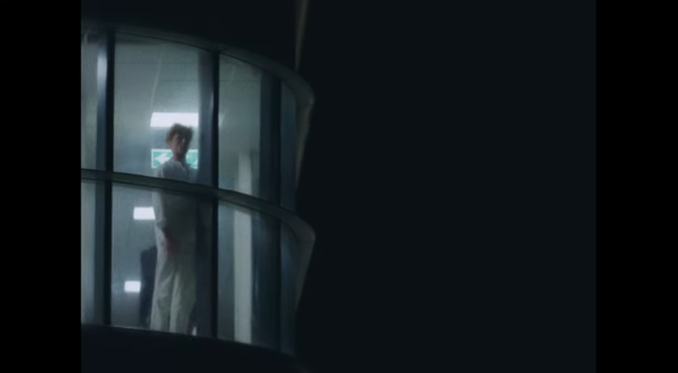 A person standing inside a curved glass elevator, viewed through the glass, with a dark background.