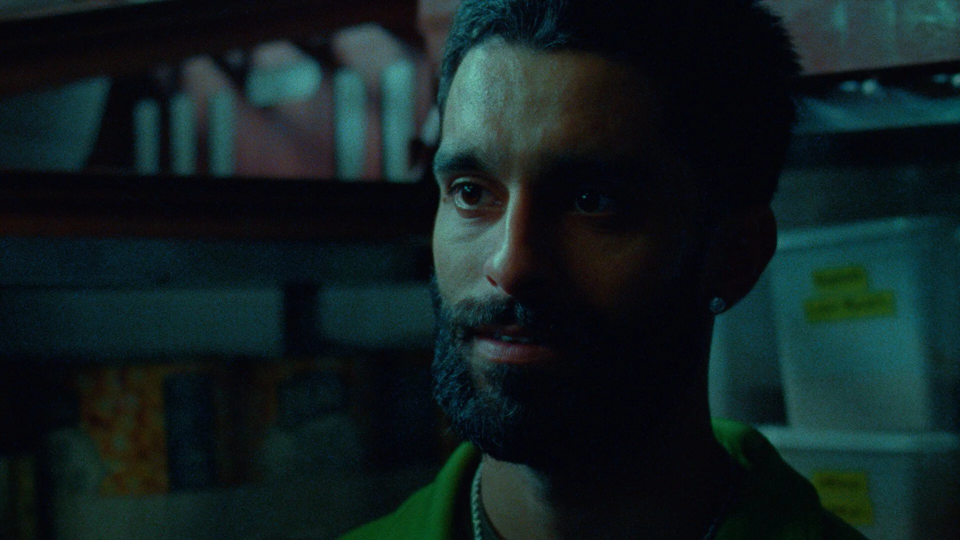 A man with dark hair and a beard looking to the side in a dimly lit room, with shelves seen in the background.