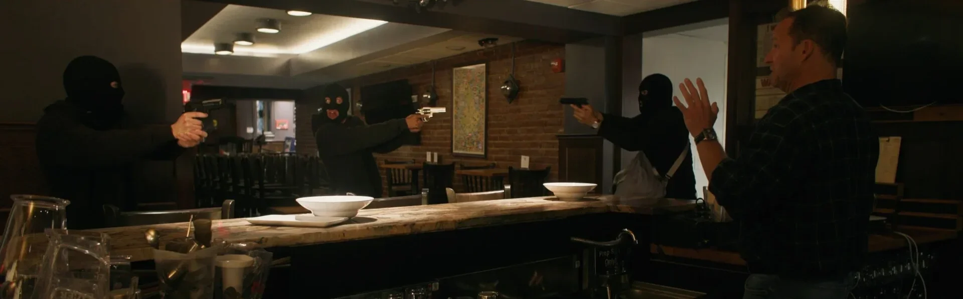 Four individuals, three wearing ski masks and holding guns, confront a man in a restaurant or bar setting. The man is raising his hands defensively.