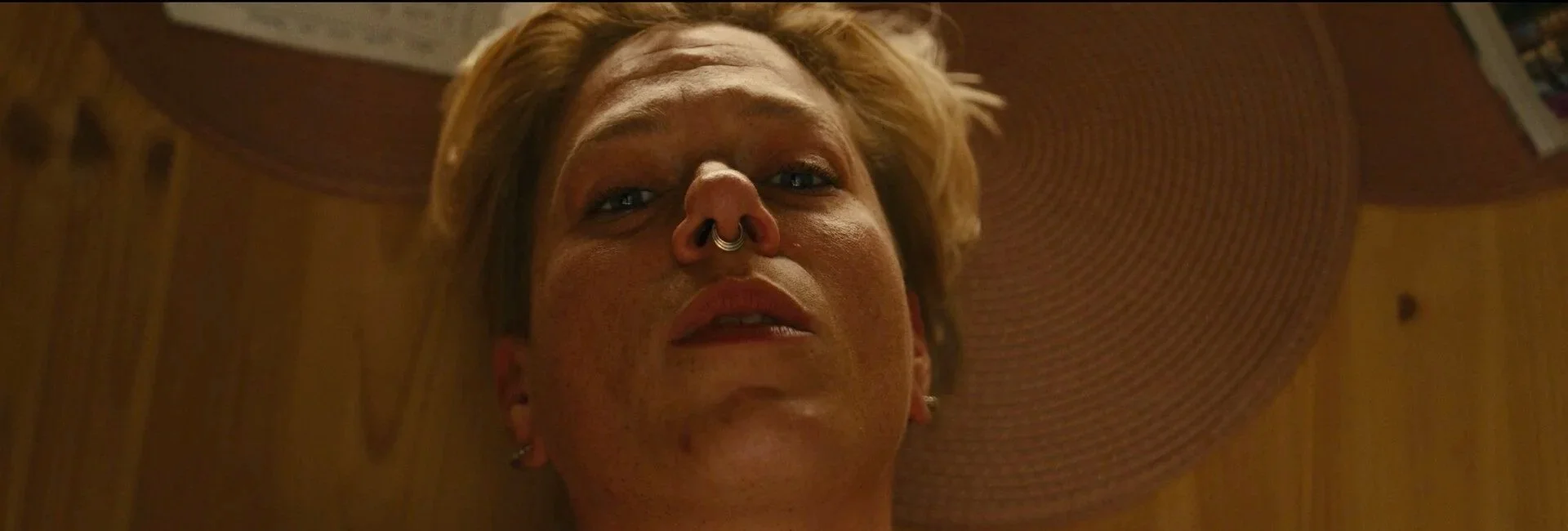 Close-up of a person's face looking down at the camera, with a septum piercing and earrings, and a wooden ceiling in the background.