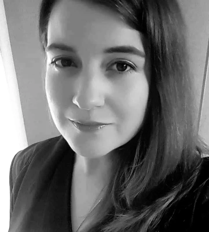 A black and white close-up photo of a woman with shoulder-length dark hair, smiling gently, wearing a dark top, standing indoors.