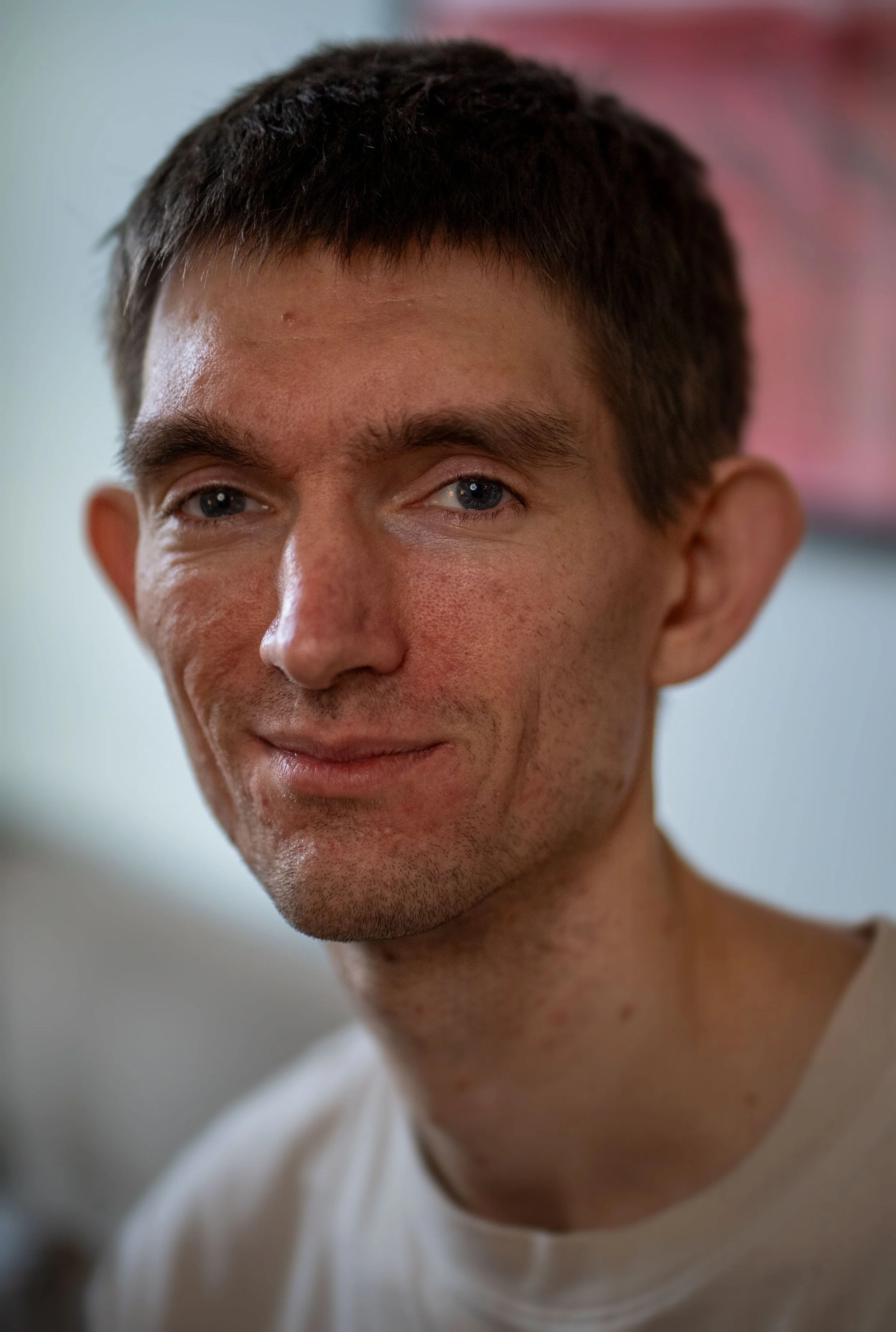 Close-up of a man with short dark hair, blue eyes, and light skin, smiling slightly, wearing a light-colored shirt, with a blurred indoor background.