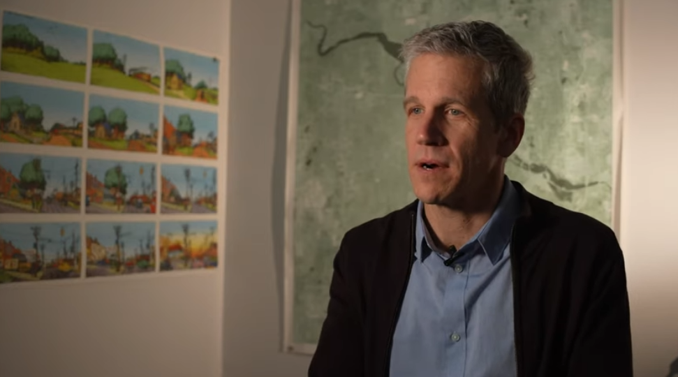 A man sitting indoors with a wall of small colorful landscape paintings and a large map behind him.