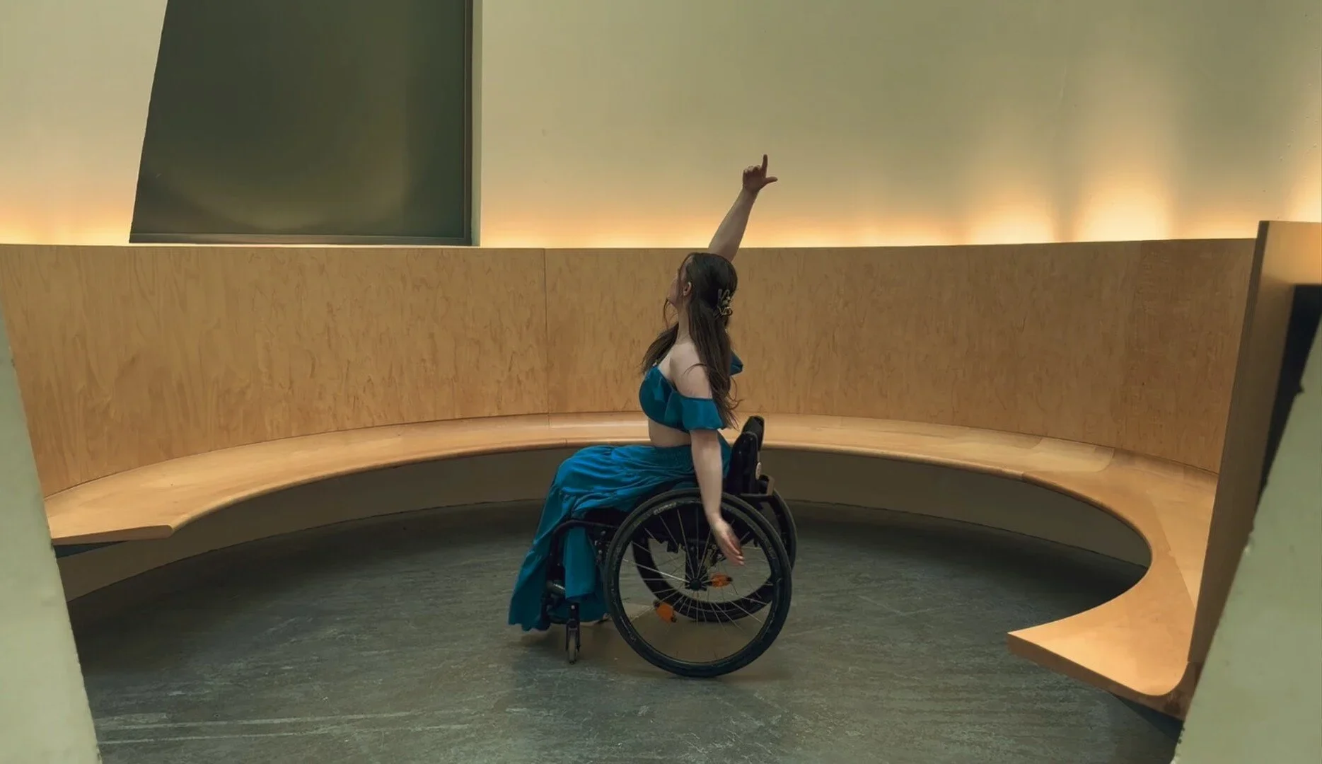 A woman in a blue dress seated in a wheelchair, performing a dance pose with one arm raised and the other arm extended downward, in an indoor setting with a curved wooden bench and a plain wall background.