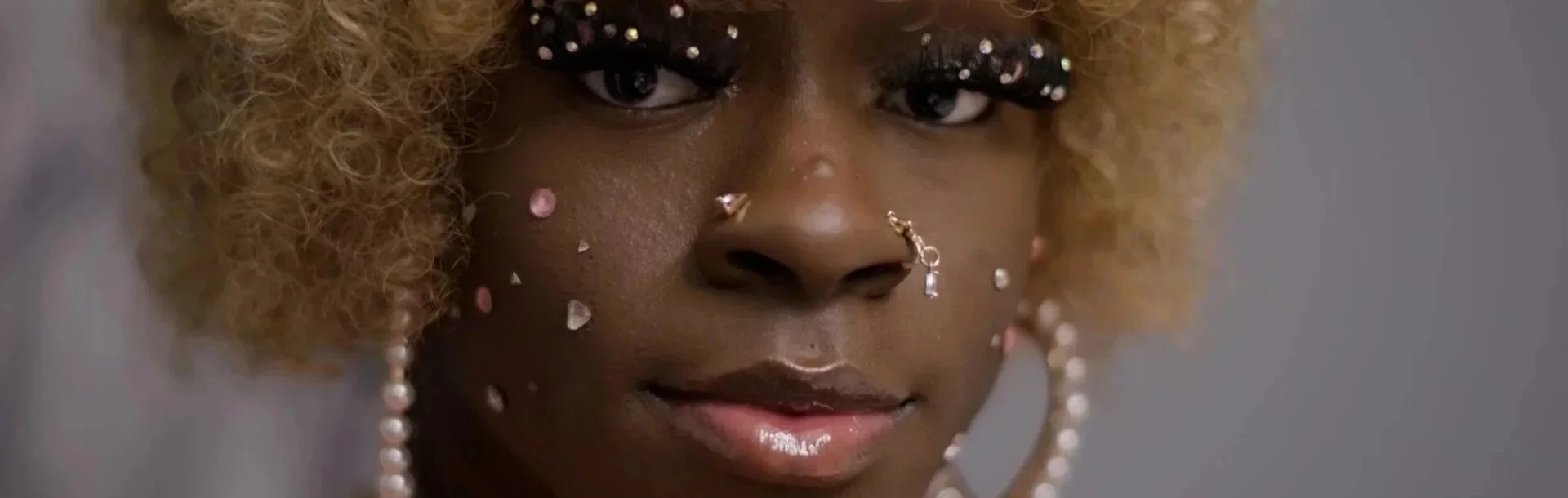 Close-up of a woman with curly blonde hair, adorned with rhinestones and jewelry on her face, wearing eye makeup with false lashes, looking directly at the camera.