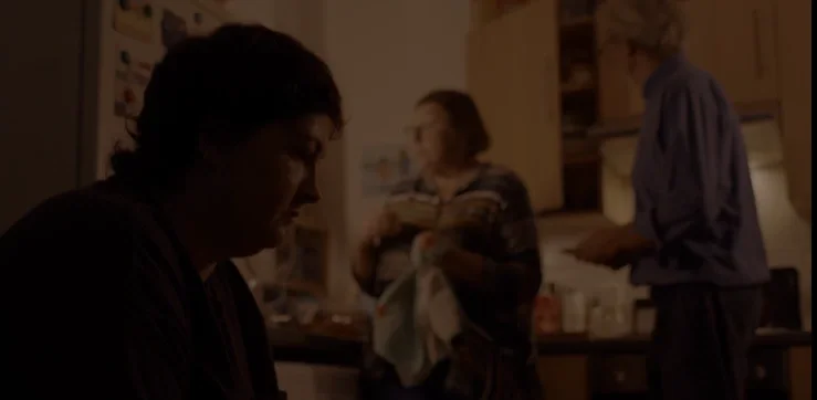 Three people in a kitchen, one sitting with head down, two standing and talking.