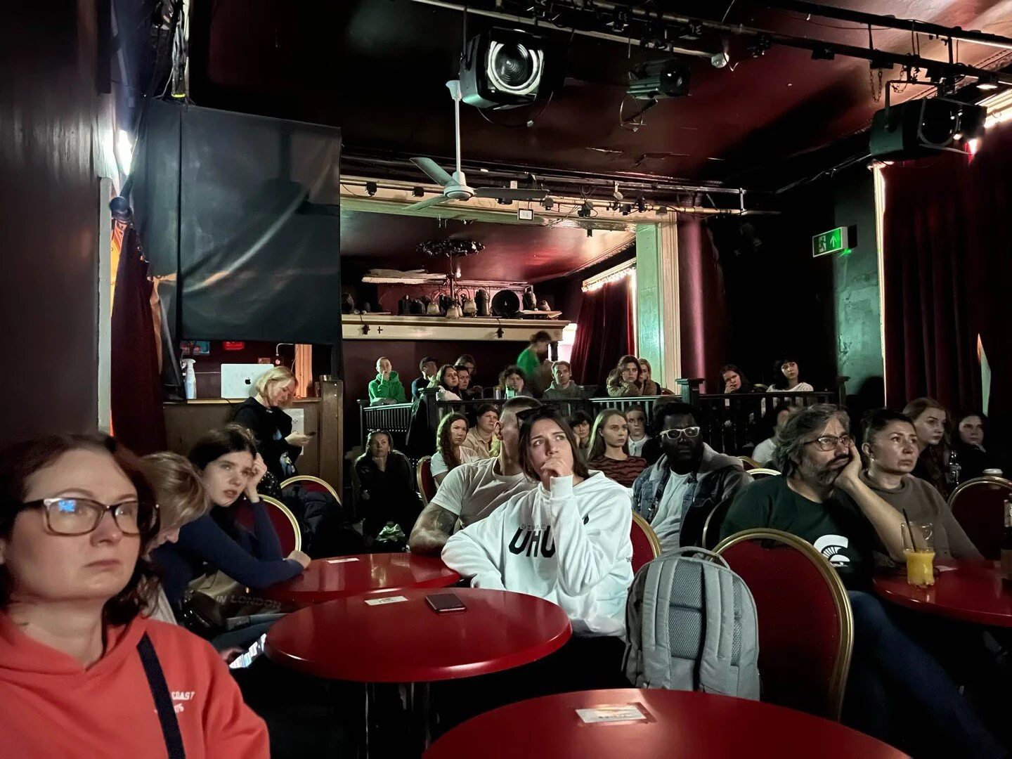 People seated at round tables inside a dimly lit comedy club or theater, watching a performance on stage, with dark red curtains and focused lighting.