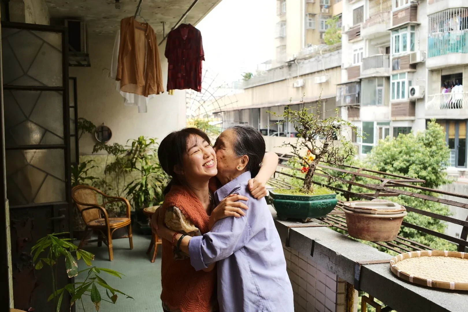 Two women hugging and smiling on an apartment balcony, with plants and hanging clothes in the background.