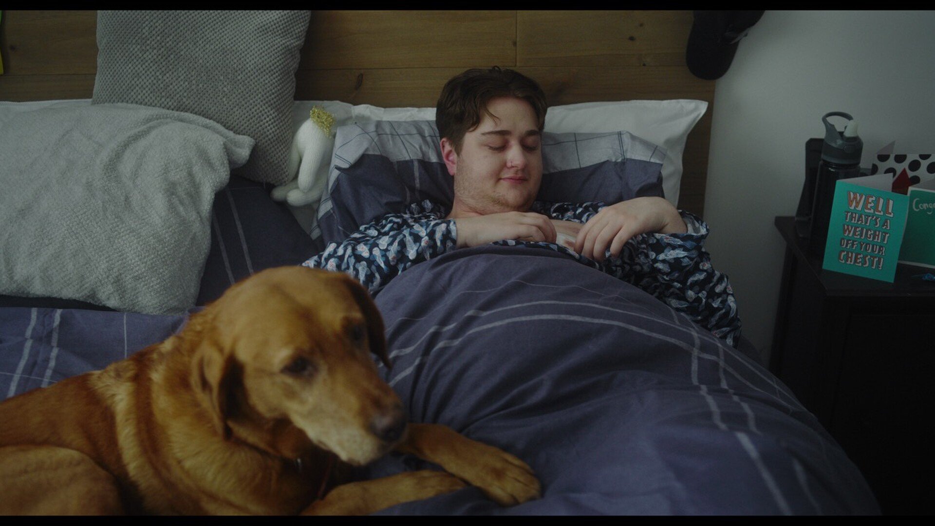 A young man is sleeping in a bed with a dog lying nearby. The room has pillows, a bedside table with a water bottle, and a card with the message "WELL THAT'S A WEIGHT OFF YOUR CHEST!"