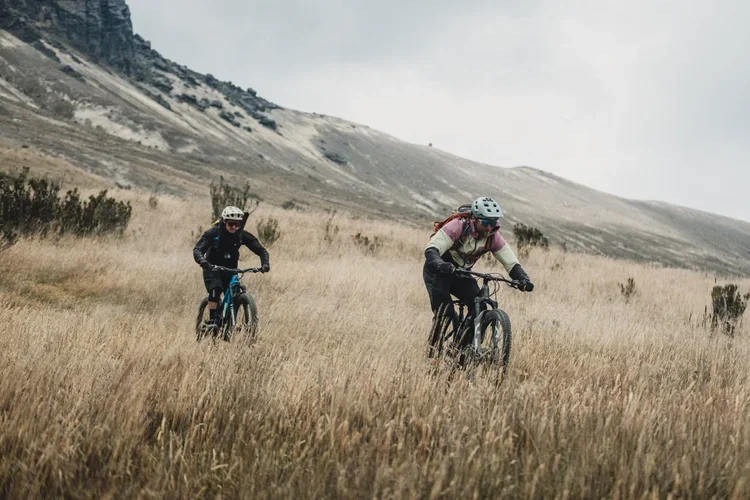 Two people mountain biking through tall grass in a hilly landscape with a cloudy sky.