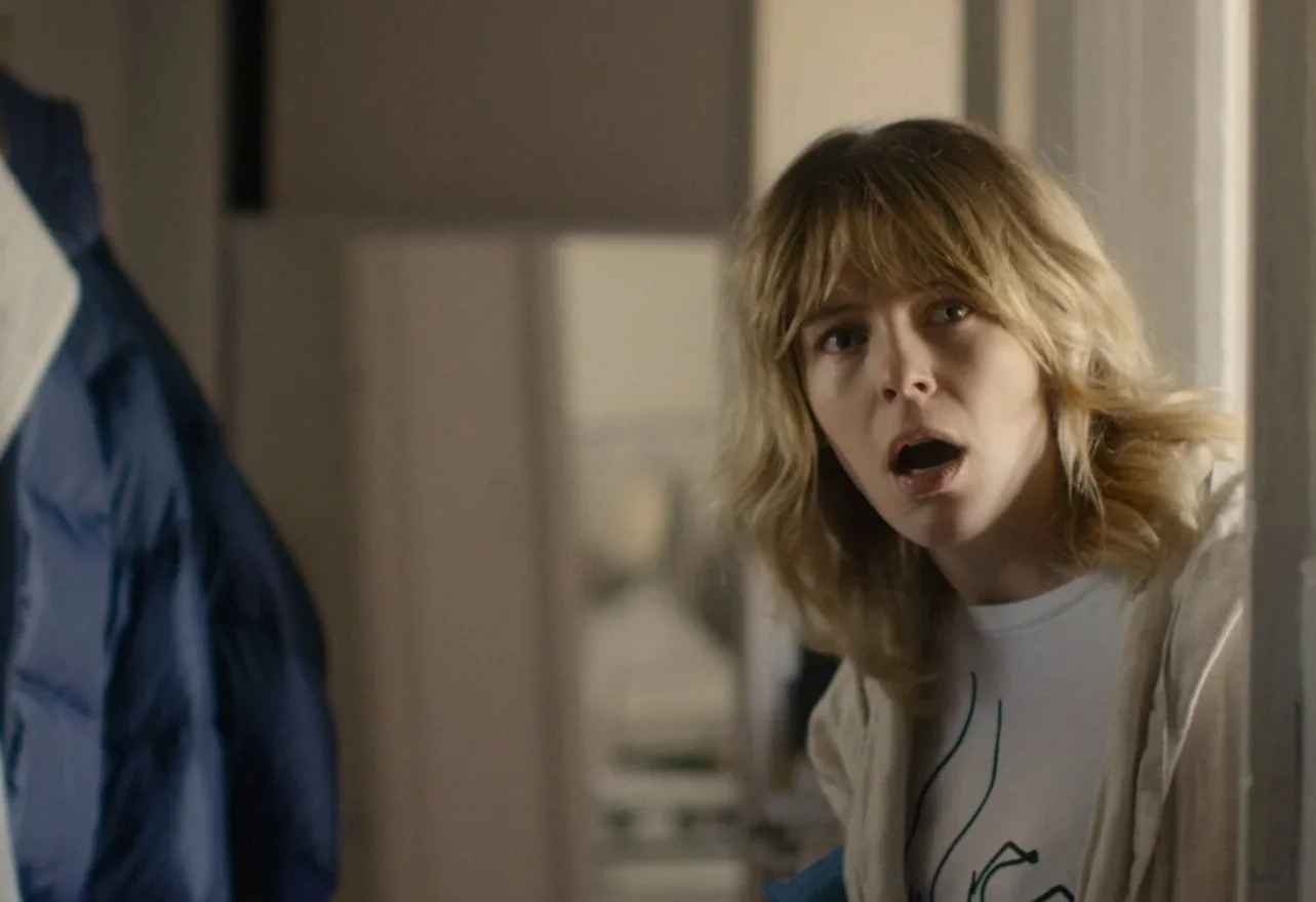 A woman with blonde, wavy hair showing a surprised or shocked expression, looking into a room or space.