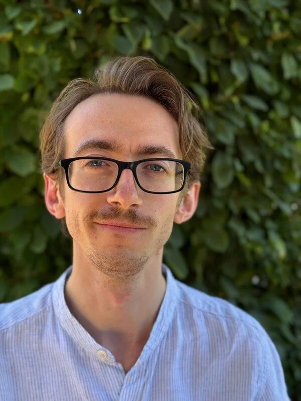 Portrait of a young man with light skin, brown hair, wearing black glasses and a light blue button-up shirt, standing outdoors in front of green foliage.