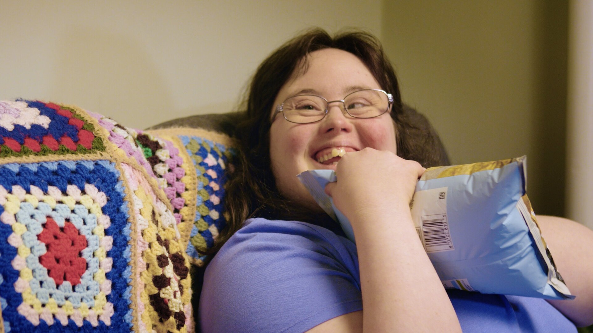 A woman with glasses, smiling, holding a bag of snacks, sitting on a sofa covered with a colorful crocheted blanket.
