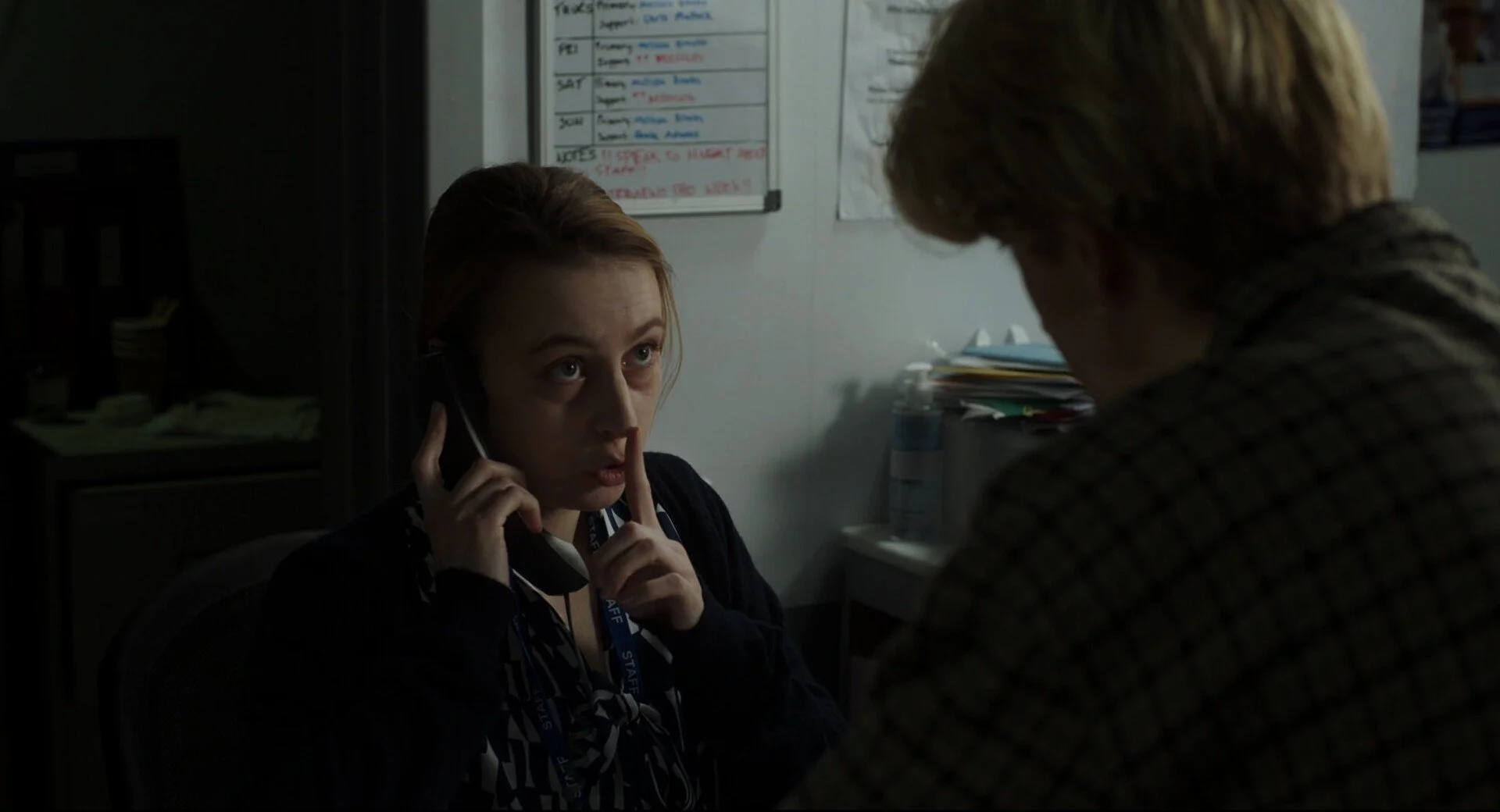 A woman with light brown hair holding a phone to her ear, with her index finger raised to her lips, in conversation with a man whose back is to the camera, in an office setting.