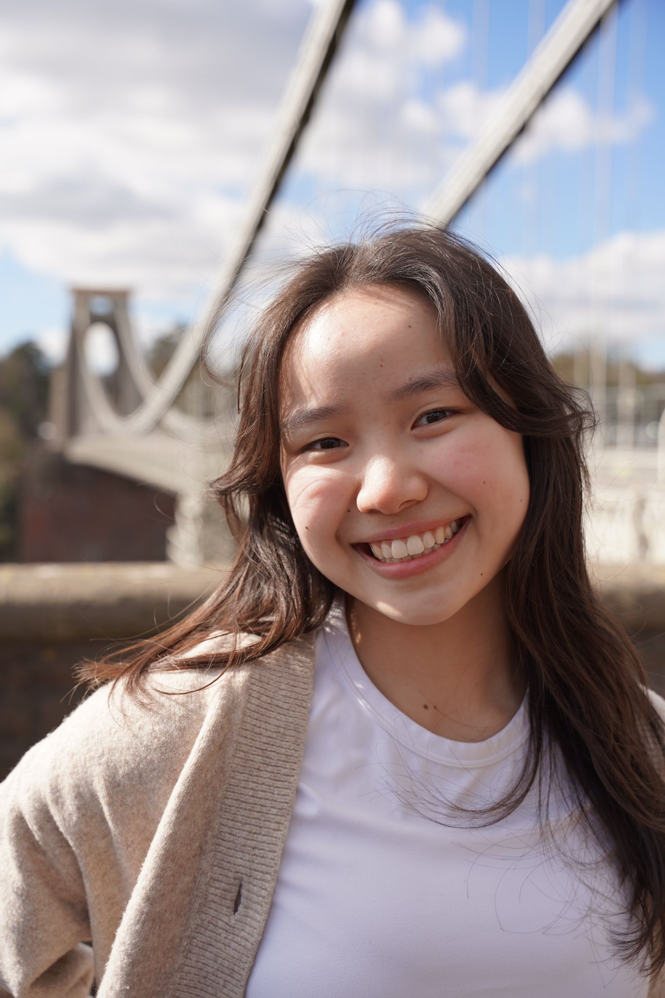 Young woman smiling outdoors near a bridge with blue sky and clouds in the background.