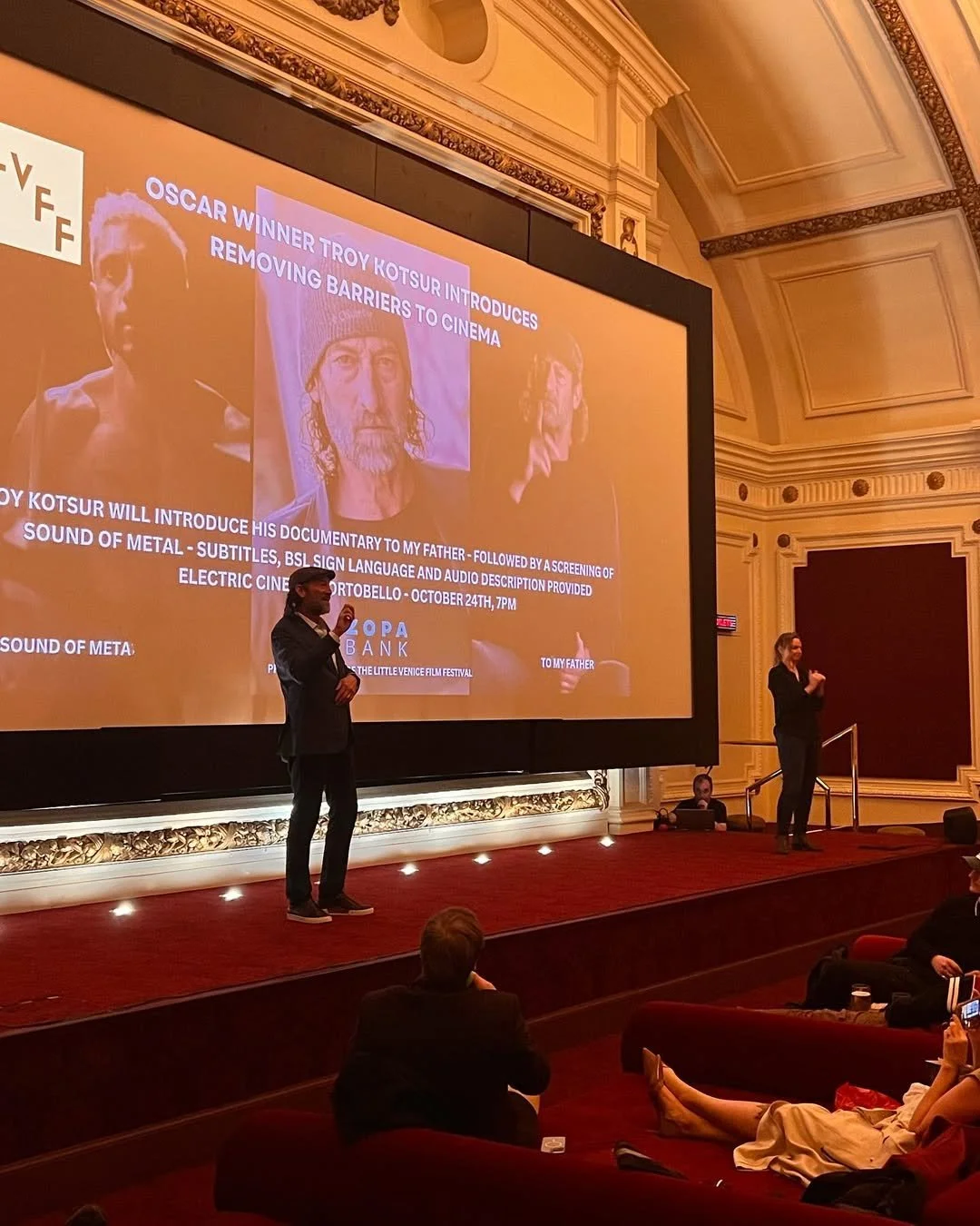 People presenting a film festival event on stage in an ornate theater. A large screen displays information about a documentary by Oscar winner Troy Kotsur, with images of other people and text about the screening.