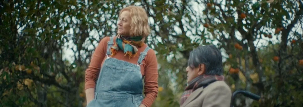 Two women in a garden or orchard, with one standing and the other sitting in a wheelchair, surrounded by fruit trees.