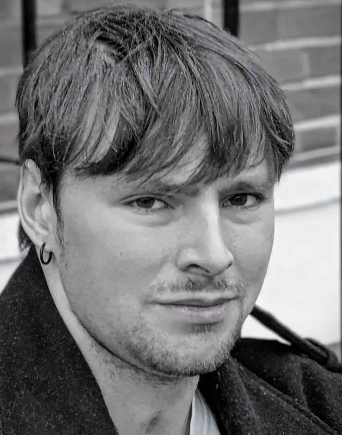 A black and white close-up image of a young man with short, tousled hair, an earring in his ear, and a faint smile, standing outdoors with a brick wall in the background.