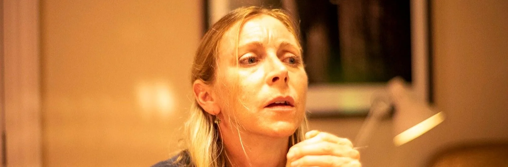 A woman with blonde hair appears emotional, with her hands clasped near her face, sitting in a dimly lit room.