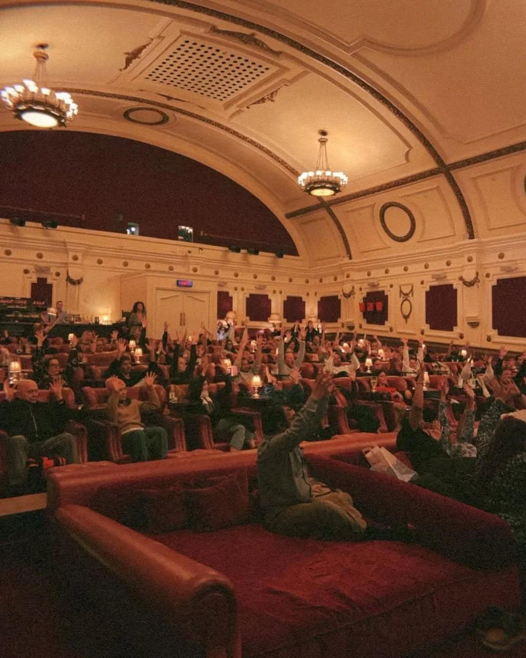 Audience in a theater with high arched ceiling, ornate chandelier, and people raising their hands.