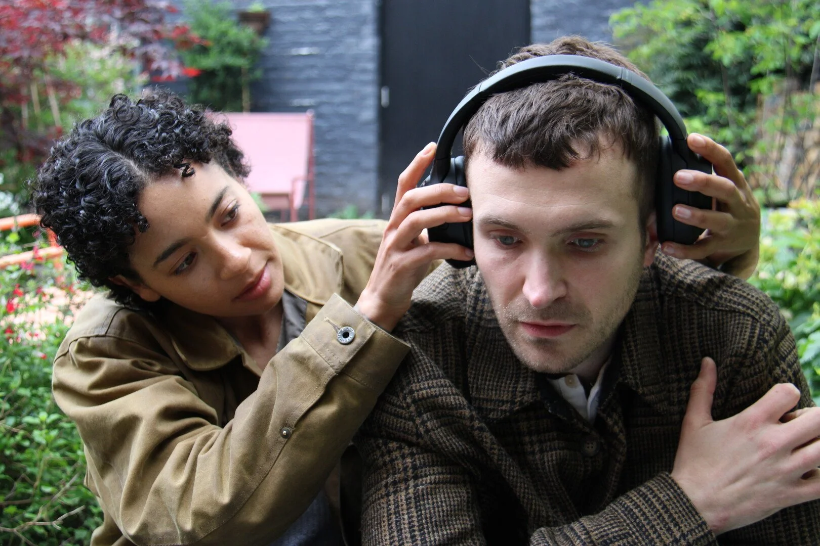A woman assists a man in adjusting his headphones in an outdoor garden setting with greenery and outdoor furniture.