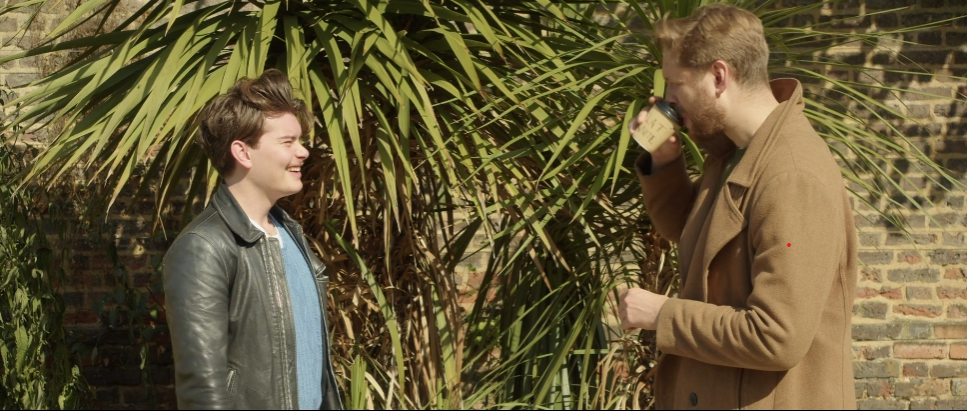 Two young men standing outdoors in front of a large leafy plant, engaging in conversation with friendly expressions. One is drinking from a mug, the other is smiling.