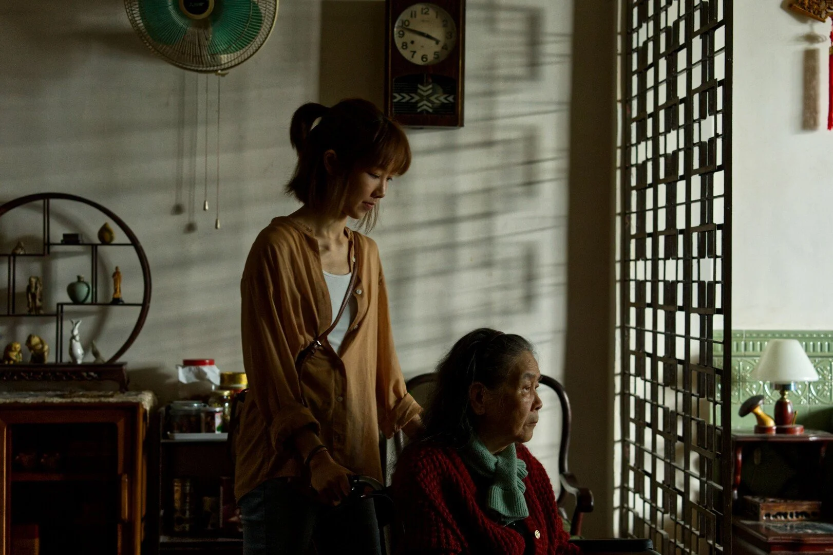 A young woman standing next to an elderly woman in a room with traditional Asian decor, sun casting shadows through a lattice screen.