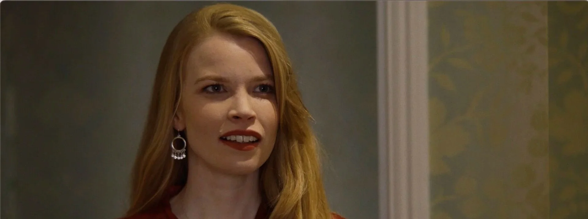 A woman with long red hair, wearing large earrings, looks confused or surprised, standing indoors against a wall with patterned wallpaper.