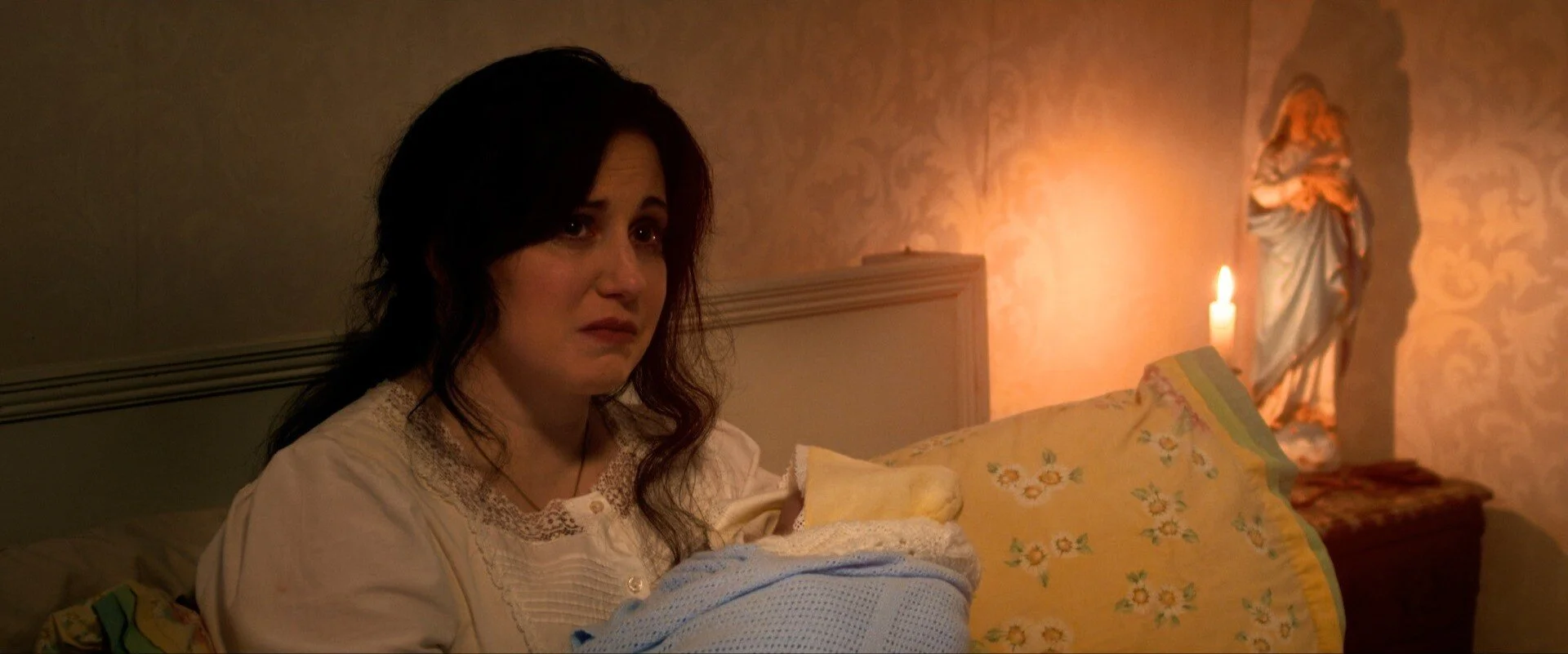 A woman with dark wavy hair sitting on a bed, looking worried, in a dimly lit room with a candle and a statue of a saint in the background.